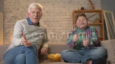 祖父和孙子坐在沙发上看电视，吃薯片，享受胜利，粉丝，快乐，微笑
